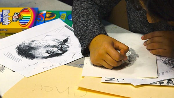 Student sketches a face in pencil sitting at a table in a classroom.