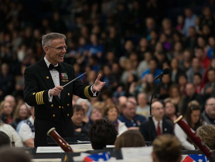 Music conductor leading the U.S. Navy Band with audience blurred behind him.