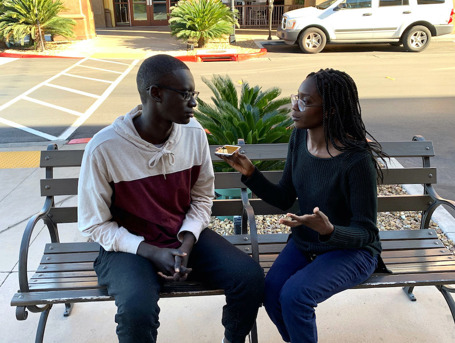 Two people sit on a bench outside. The woman on the right records the man on the left with her phone.