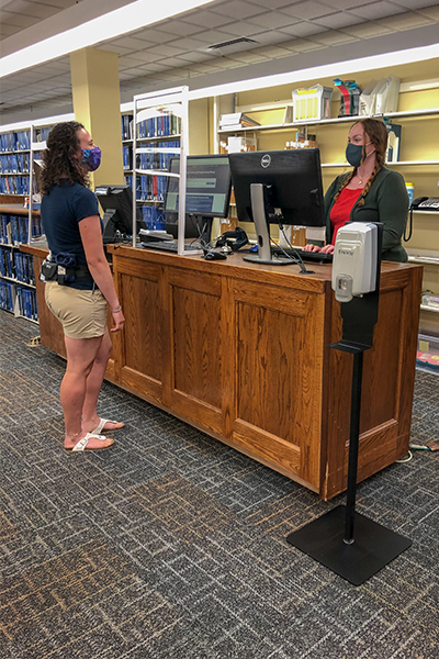 A dark-haired student wearing a face covering stands at the DeLaMare Circulation desk as a DeLaMare staff member wearing a face covering retreives the student's checked-out library materials.