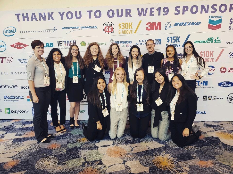 A group of SWE students pose for a photo together in front of a wall with sponsor names on it. 