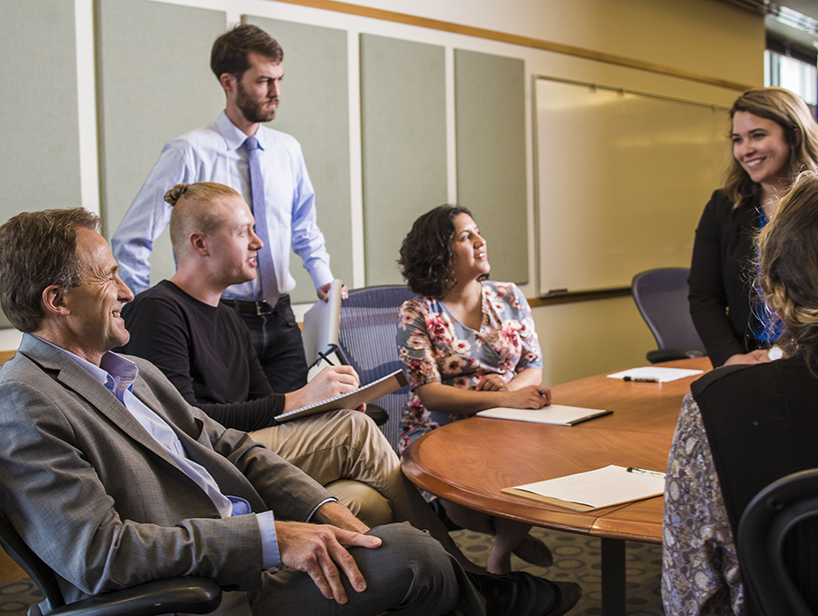 Six people sit around a conference table in a bright room smiling and talking.