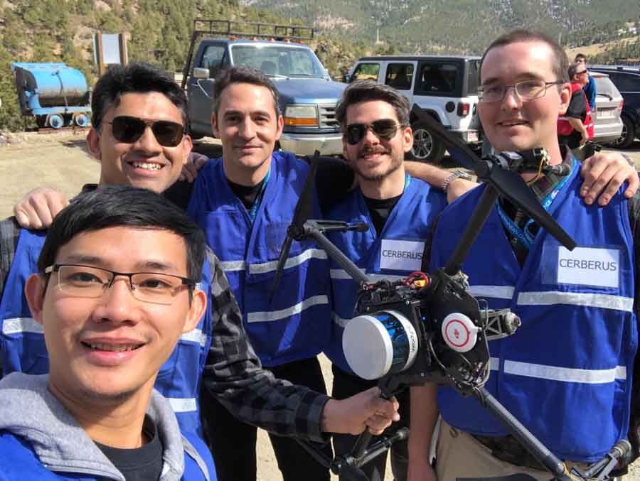 A group of researchers in blue vests are posing with one of their robots during the DARPA competition. 