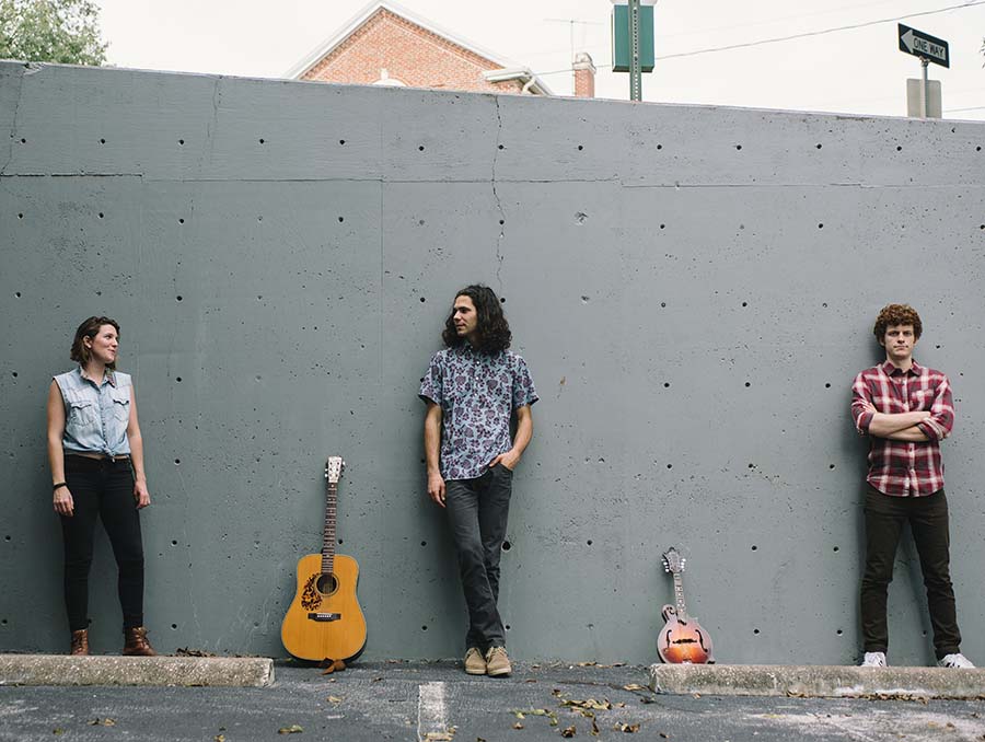 Three members of the Steel Betty blue grass group stand in front of an outdoor concrete wall with guitars propped up on the wall between each of them.