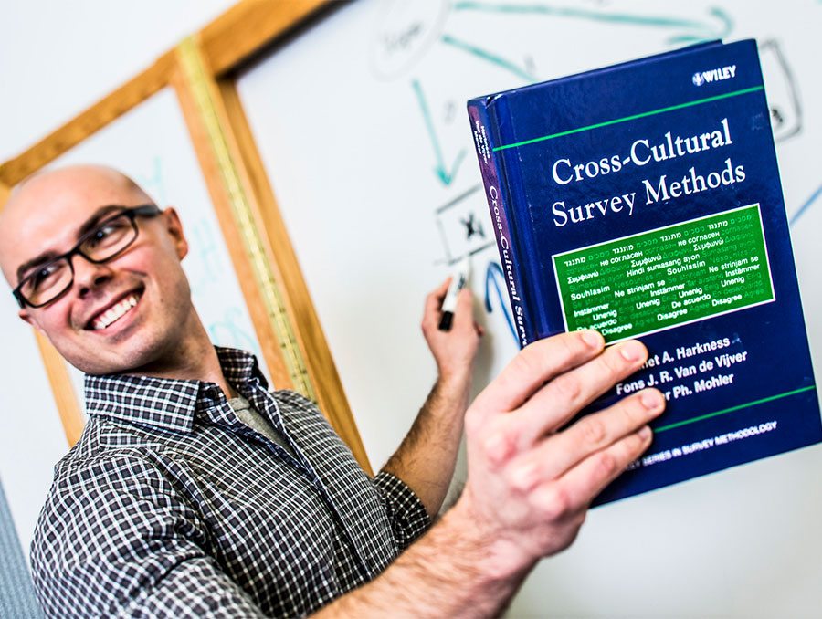 Instructor teaching in front of a whiteboard while holding a textbook titled, "Cross-Cultural Survey Methods" by Harkness, Van de Vijver, and Mohler. 