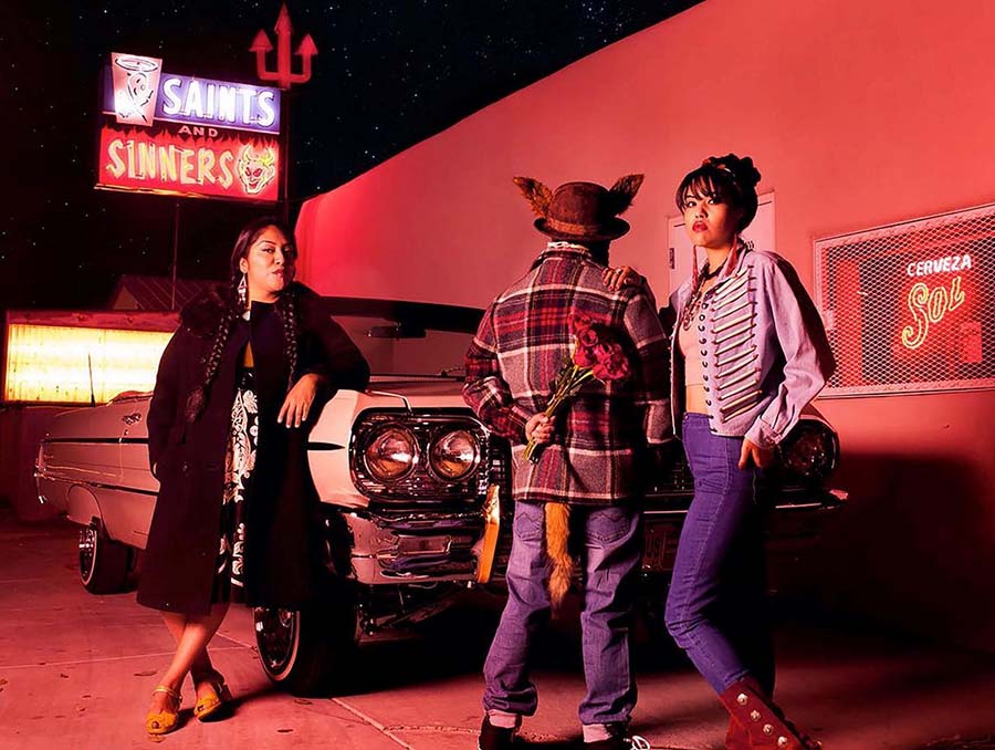 Photograph by Cara Romero titled “Coyote Tales," which includes a Saints and Sinners neon sign and three people standing outside a liquor store in front of a classic, convertible car. 