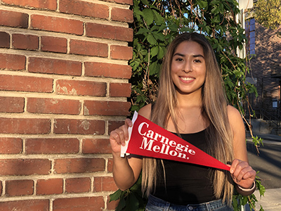McNair Scholar Valeria Nava standing in front of a brick wall holding a Carnegie Mellon flag