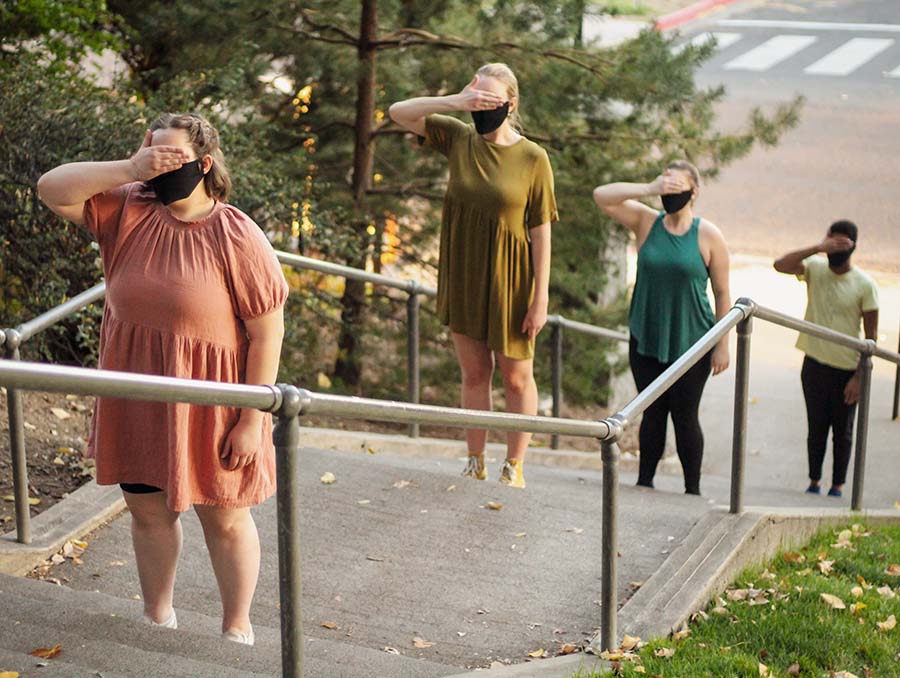 People wearing masks stand on different stairs on an outside staircase covering their eyes with their right hands.