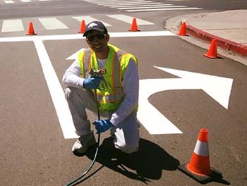 A person crouches down with a paint suit and mask to paint the street with directional arrows.