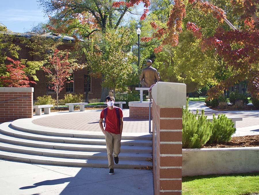 A student wear a facial covering walks through the University of Nevada, Reno campus with stairs and a statue behind them.