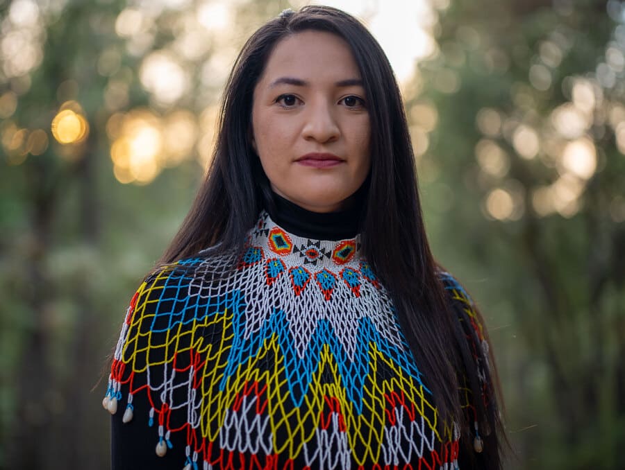 Tsanavi Spoonhunter stands in front of a forest backdrop wearing a Native American beaded collar. 