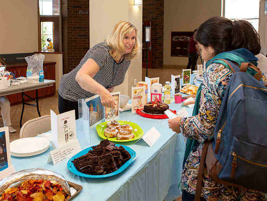 Woman in a striped shirt stands behind a table with a variety of edible baked goods. She is welcoming a University student to the Edible Books Festival inside the Mathewson-IGT Knowledge Center Breezeway. Each baked good on the table is based on a banned book.