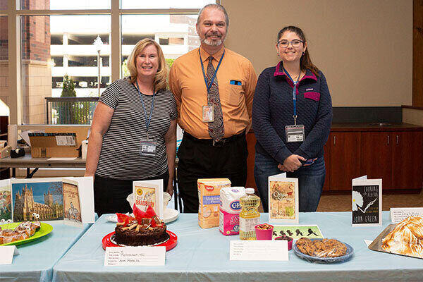 Two women and one man stand behind a table in the Breezeway of the Mathewson-IGT Knowledge Center overlooking a variety of baked goods submitted to the Libraries annual Edible Book Festival event, which is part of Banned Books Week.