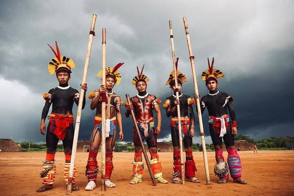 A group of men stand in front of a cloudy sky holding spears and wearing body paint.