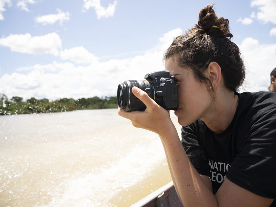 Luiza Vieira holds a camera to her face in front of a beach with greenery and the sky in the backdrop.