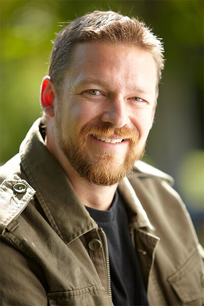 Photo of man with light-colored hair and beard wearing a tan jacket and black shirt 