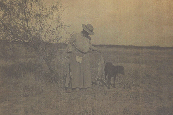 Scanned image of a woman holding a rifle and standing next to a black dog.