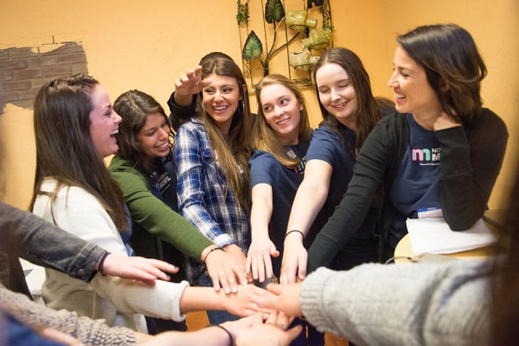 Students and a professor pose indoors, putting their hands in a circle