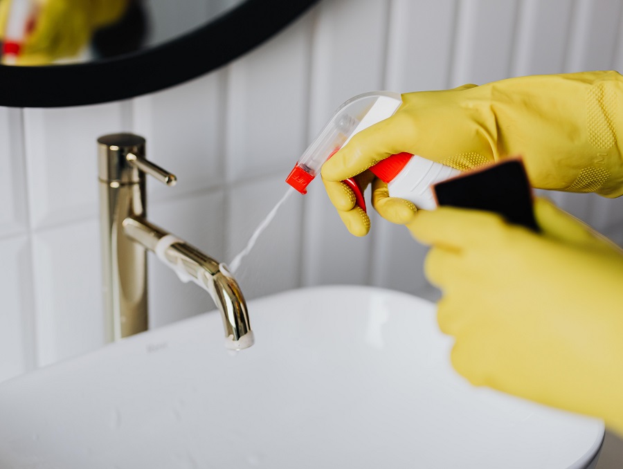 Hands in yellow cleaning gloves spraying disinfectant on a bathroom faucet