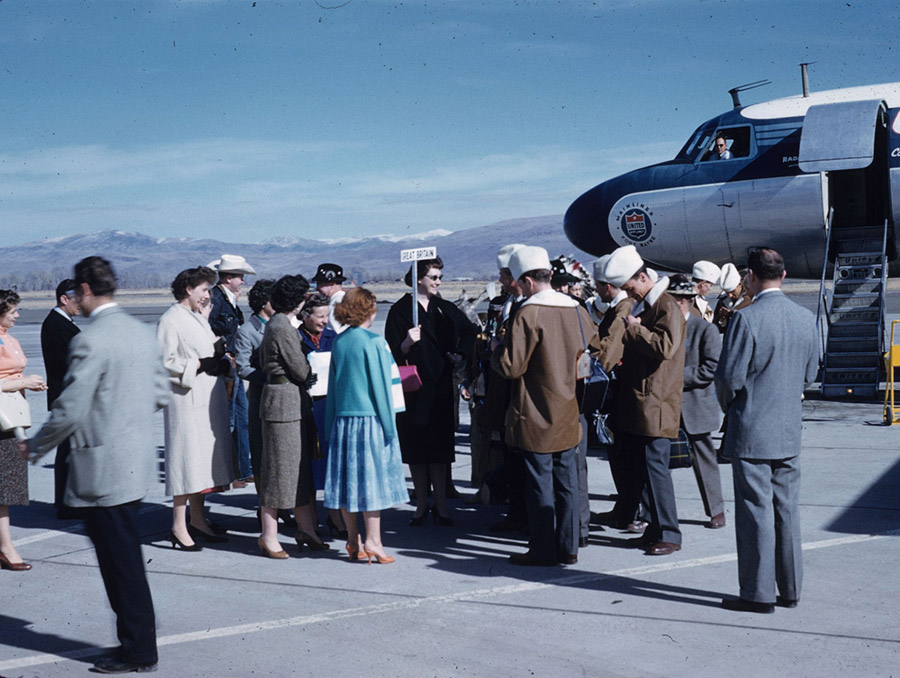 A color photograph showing a crowd standing on the tarmac at the Reno Tahoe Airport. A woman holds a sign with “Great Britain” on it, while a group of people wearing matching brown coats with white hats stand near her. A United Airlines airplane is in the background with a stairway down to the tarmac.