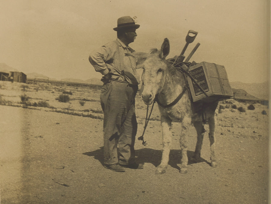 Sepia-toned photograph of a man standing next to a donkey.