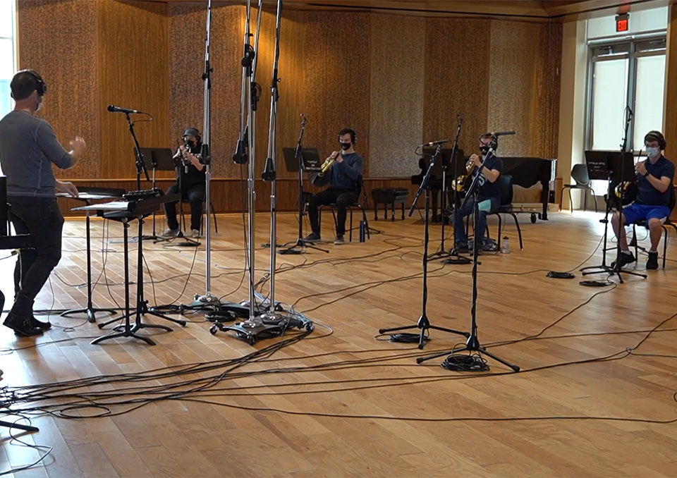 Student musicians sit socially-distanced in band room and record song with conductor