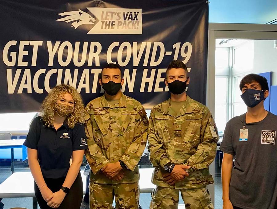 Four people in masks stand in front of a banner reading, "Get your COVID-19 vaccination here."