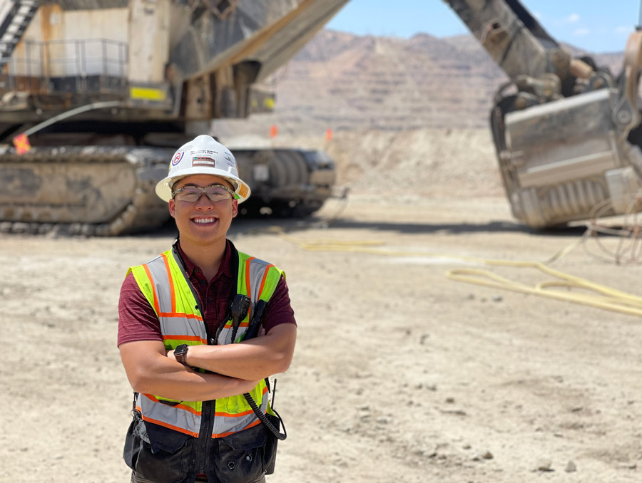 Brian Huynh, wearing a white work helmet and a reflective vest, stands smiling with his arms crossed in the foreground of a mine site.