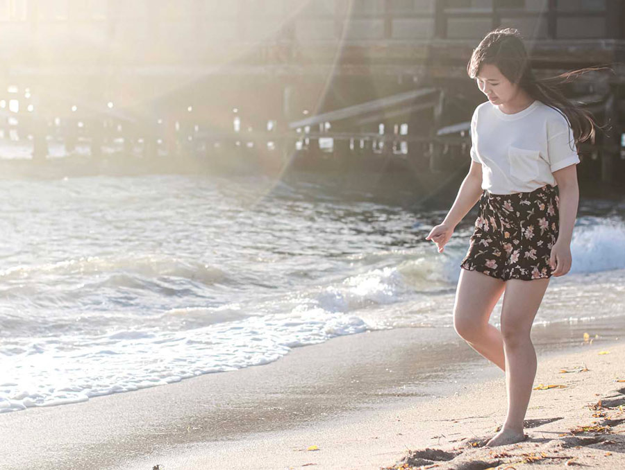A girl with dark hair, a white shirt and a floral print skirt walks along a beach near a pier.