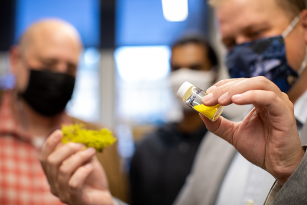A masked Christopher Jeffrey holds a vial containing a bright yellow crystalline powder labeled "vulpinic acid" in one hand and a blurry green lichen in the other, with two other individuals in the background.