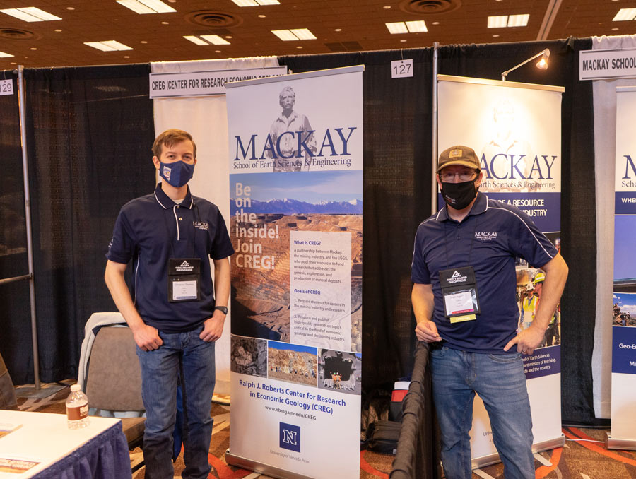 Two people each stand in front of a booth display for CREG and the Mackay School.