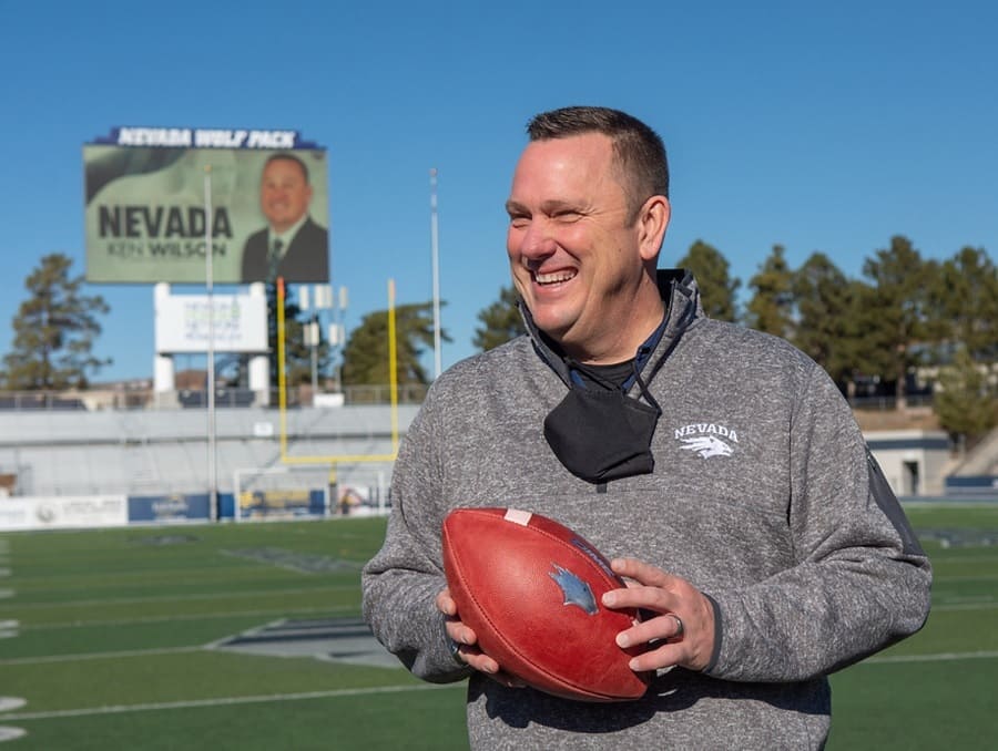 Ken Wilson holding a football on the Mackay Stadium field with a large screen announcing his new role behind him