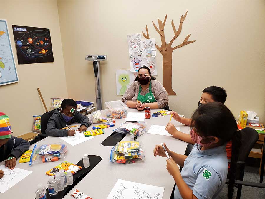 Samantha Shoupe and 4-H youth sit around a long rectangular table covered in craft supplies as they each draw reindeer. Educational posters (including one of the solar system) and kids' artwork adorn the walls.