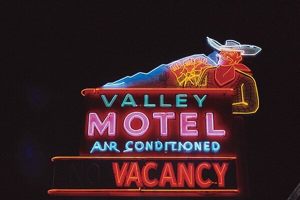 Colorful neon motel sign featuring the neon outline of a cowboy with the outline of white neon mountains in the background. The sign reads Valley Motel Air Conditioned VACANCY