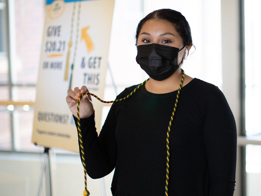 A student wearing a mask holds up a gold and green graduation tassel which hangs around her neck.