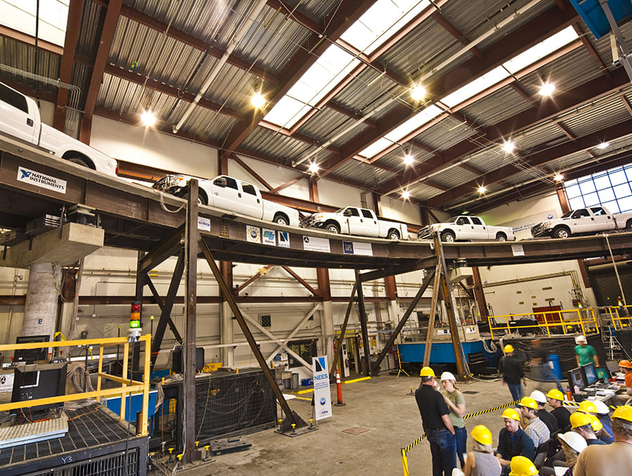 Six trucks on curved bridge spanning four shake tables with onlookers viewing experiment