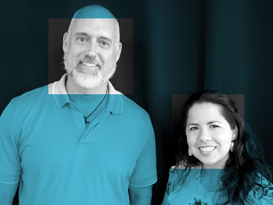 A man and a woman post on a black backdrop. The image is gray scale with a blue filter over the top. The filter is removed on both of the faces of the man and woman. 