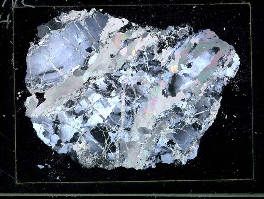 Thin section of brecciated quartz appears on a black background. Colors in the image appear as silvery, blue tones with flecks of black throughout