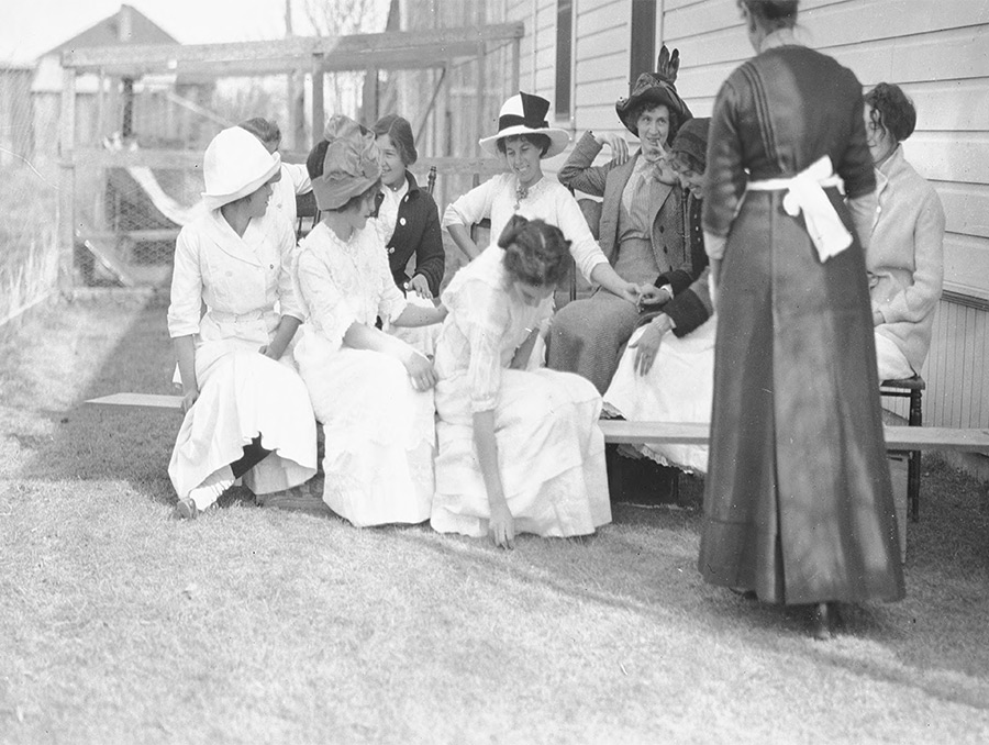 black and white photograph of a group of women seated outside; one women wearing apron with back turned to camera.