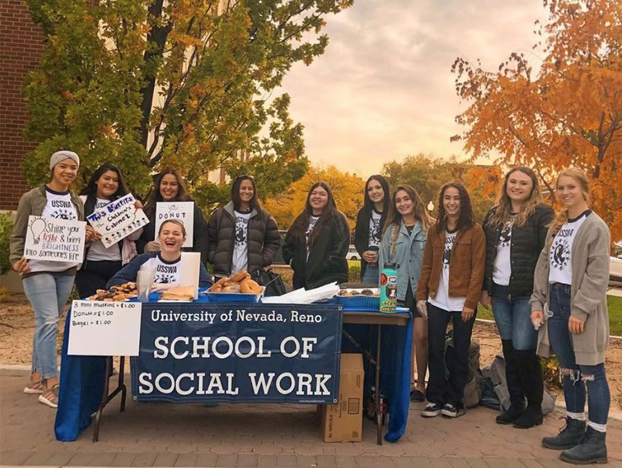 A group of University students with the School of Social Work stand around a table with signs and donuts