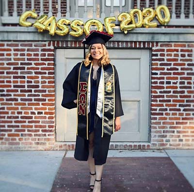 Class of 2020 gold balloons hung outside of a building with a graduating student standing in front of building