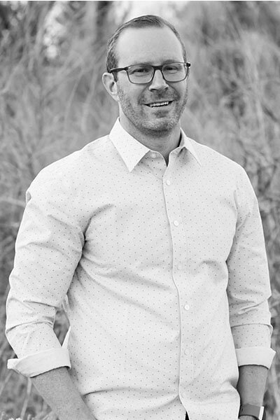 Black and white photo of a man wearing glasses and a light-colored long sleeve shirt standing outside in a field.