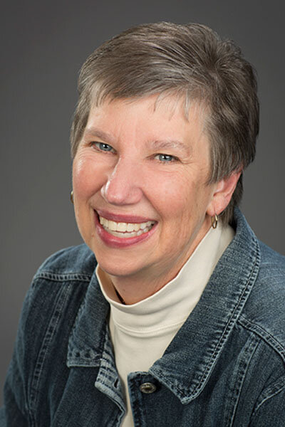 Photo of a woman with short, grey hair wearing a white turtle neck, dark denim jacket and small hoop earrings.