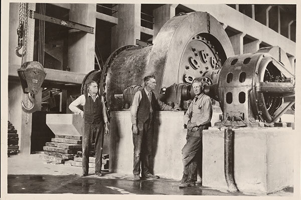 Three men stand in front of large copper processing machinery twice their size