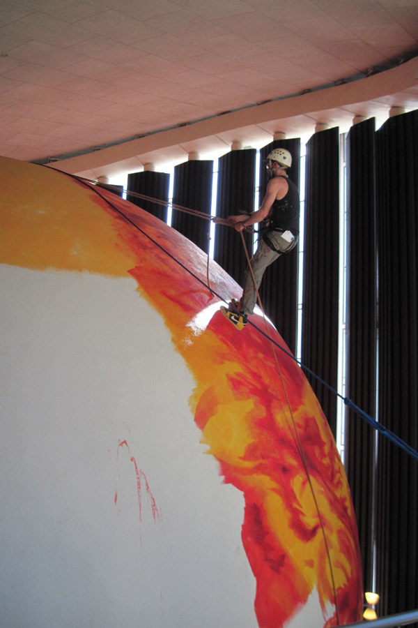 A man works on repainting the dome theater, suspended by ropes.