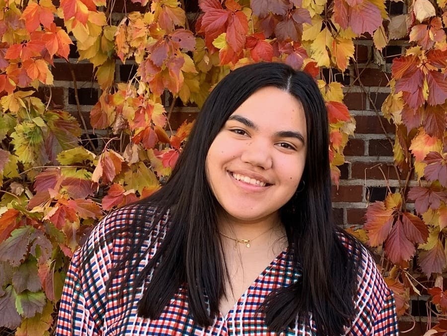 Ashlyn Rodgers smiles in front of a brick wall that is covered with autumnal leaves