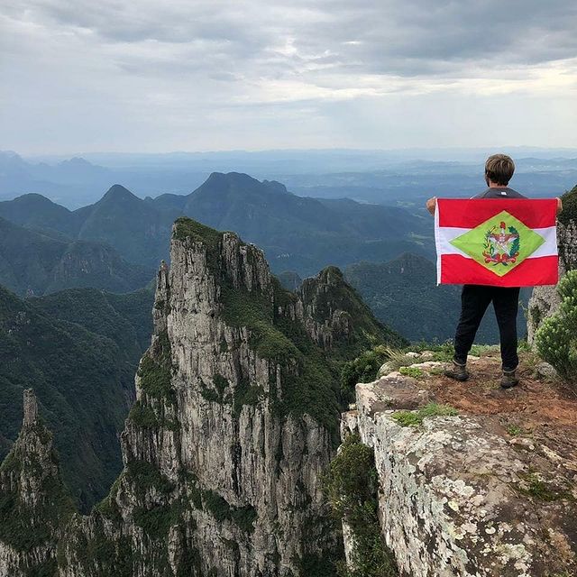 Connor with his flag-draped back to the camera stands on a high ledge and looks out over a view of towering, rocky, tree-topped cliffs and mountains.