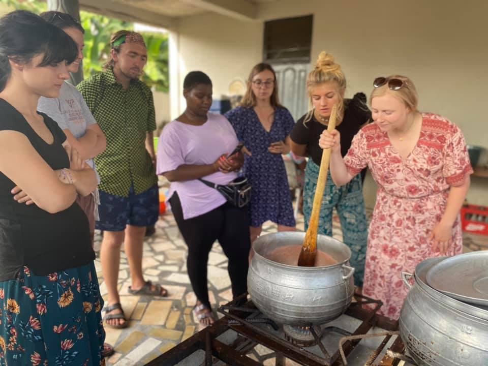 Connor Billman and five fellows watch curiously as a sixth fellow stirs a huge pot of food cooking on an outdoor gas stove with several-foot-long wooden spoon.