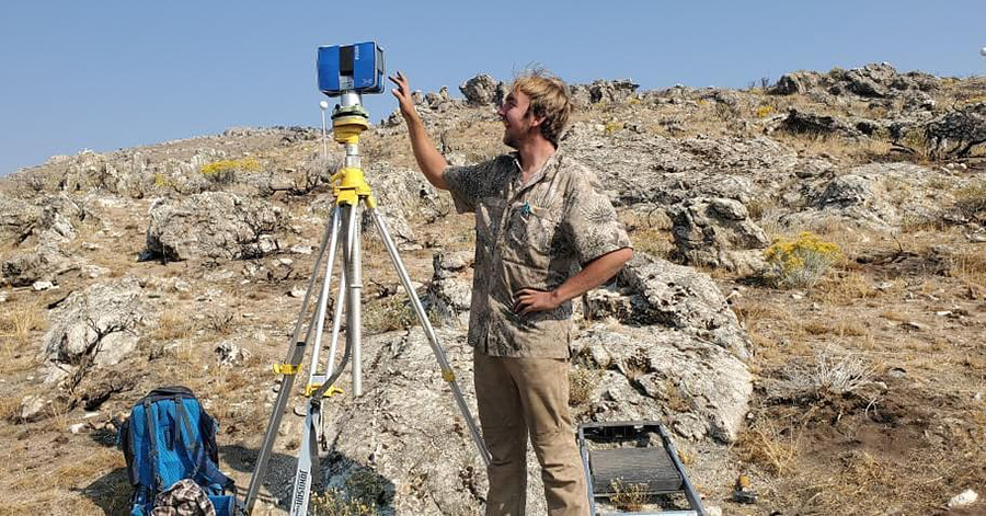 Connor on a remote rengeland, setting up a tall silver tripod upon which is a blue box with white antenna. At the base of the tripod is his backpack and a ladder.