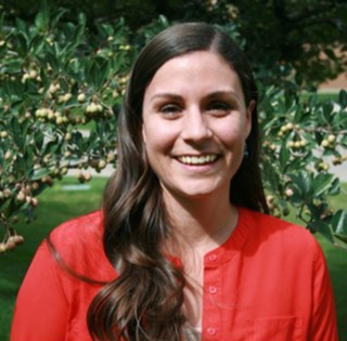 Bethany Contreras smiles, wearing a red shirt and standing in front of green foliage.